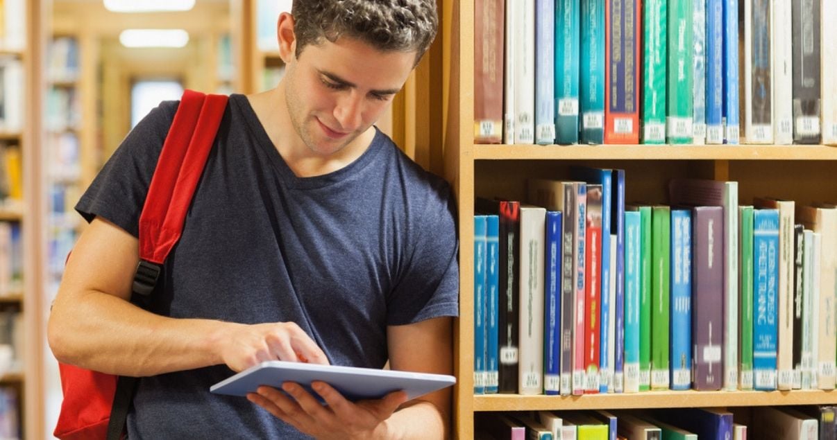 Joven con ipad en una biblioteca aprendiendo sobre los verbos irregulares.