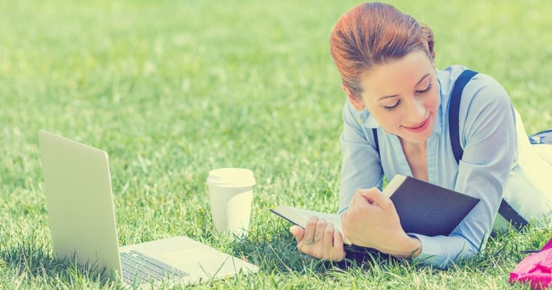 Mujer joven sonriendo acostada sobre el pasto con su laptop y un libro estudiando los verbos regulares e irregulares en inglés