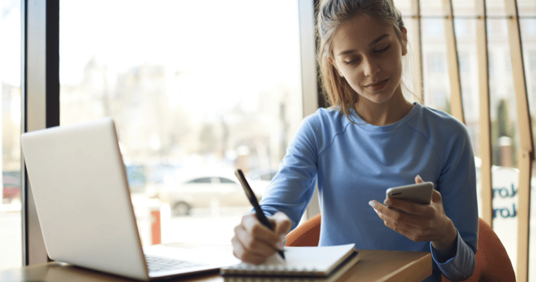 Una mujer joven anotando slangs con su teléfono y la computadora para poder adquirir más vocabulario en inglés